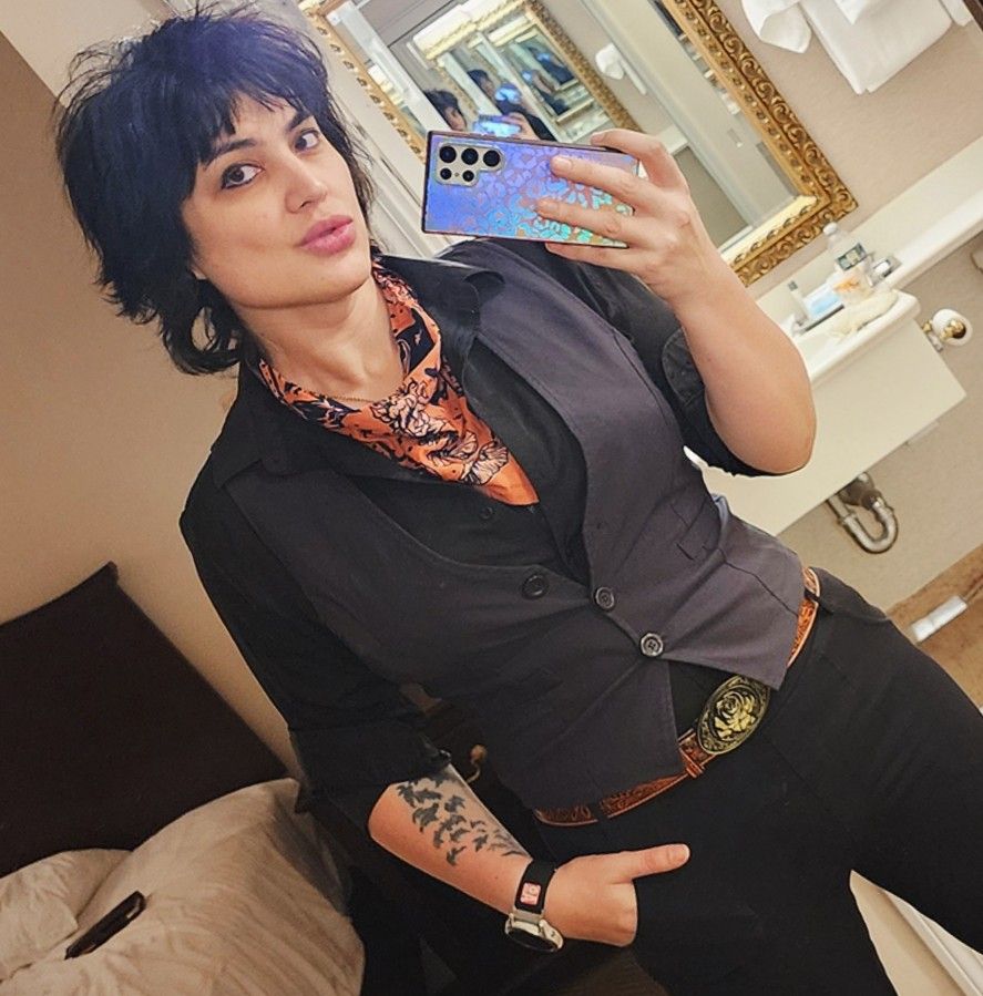 A mid-30's latina with a wolf cut hair style faces the camera. She's dressed in black shirt, a waistcoat, a belt buckle, and she has a hankerchief around her neck. She has tattoos on her right arm.